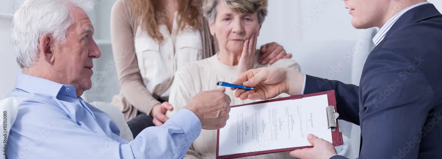 Ein Mitarbeiter von Engel Behördenservice übergibt einem alten Mann den Stift für die Unterzeichnung seines Testaments, während eine besorgte alte Dame von einer Frau getröstet wird
